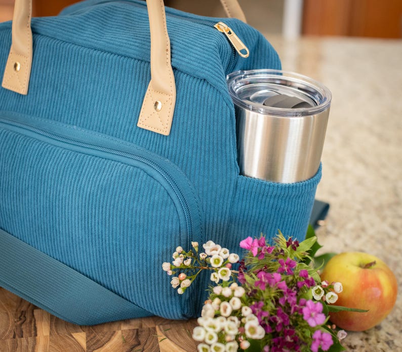 May include: Teal corduroy lunch bag with tan leather handles and a gold zipper. A stainless steel tumbler sits in the side pocket. A small bouquet of flowers and a yellow apple are next to the bag.