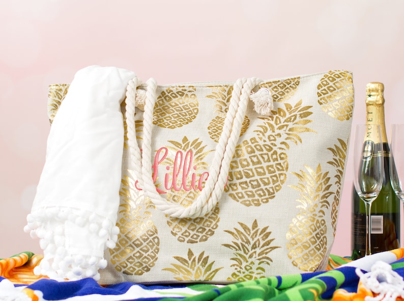 May include: A white canvas tote bag with gold pineapple print and rope handles. The bag is personalized with the name "Lillie" in pink lettering. A white towel with pom-poms is in the bag.