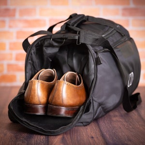 K&ouml;nnte beinhalten: Eine schwarze Reisetasche mit Rei&szlig;verschluss und einem braunen Ledergriff. Zwei braune Lederschuhe befinden sich in der Tasche.