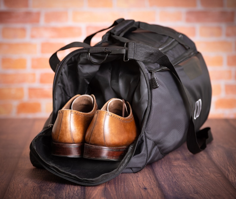 K&ouml;nnte beinhalten: Eine schwarze Reisetasche mit Rei&szlig;verschluss und einem braunen Ledergriff. Zwei braune Lederschuhe befinden sich in der Tasche.