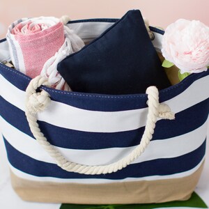 May include: A striped tote bag with navy blue and white stripes, a tan bottom, and rope handles. The bag is filled with a rolled red and white striped towel, a navy blue pillow, and a pink flower. Perfect for a day at the beach.