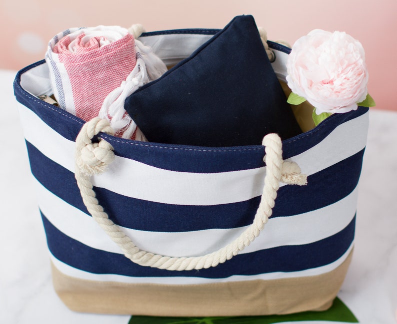 May include: A striped tote bag with navy blue and white stripes, a tan bottom, and rope handles. The bag is filled with a rolled red and white striped towel, a navy blue pillow, and a pink flower. Perfect for a day at the beach.