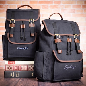 May include: Two black canvas backpacks with brown leather accents. One backpack has the name "Gianna, RN" embroidered on the front, and the other has the name "Sydney" embroidered on the front.