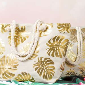 May include: Two beige canvas tote bags with gold leaf tropical leaf designs. The bags have rope handles.