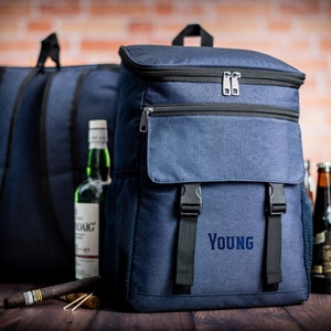 K&ouml;nnte beinhalten: Ein blauer isolierter Rucksack mit einer gro&szlig;en Fronttasche und dem Wort "YOUNG" auf der Vorderseite gestickt. Der Rucksack hat verstellbare Tr&auml;ger und einen Rei&szlig;verschluss.
