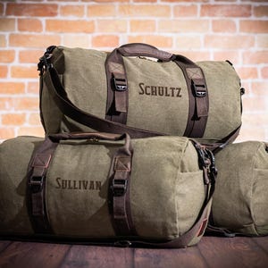 K&ouml;nnte beinhalten: Drei gr&uuml;ne Canvas-Reisetaschen mit braunen Lederakzenten und Riemen. Die Taschen sind mit den Namen "Schultz", "Sullivan" und einem dritten Namen personalisiert, der auf dem Bild nicht zu sehen ist.