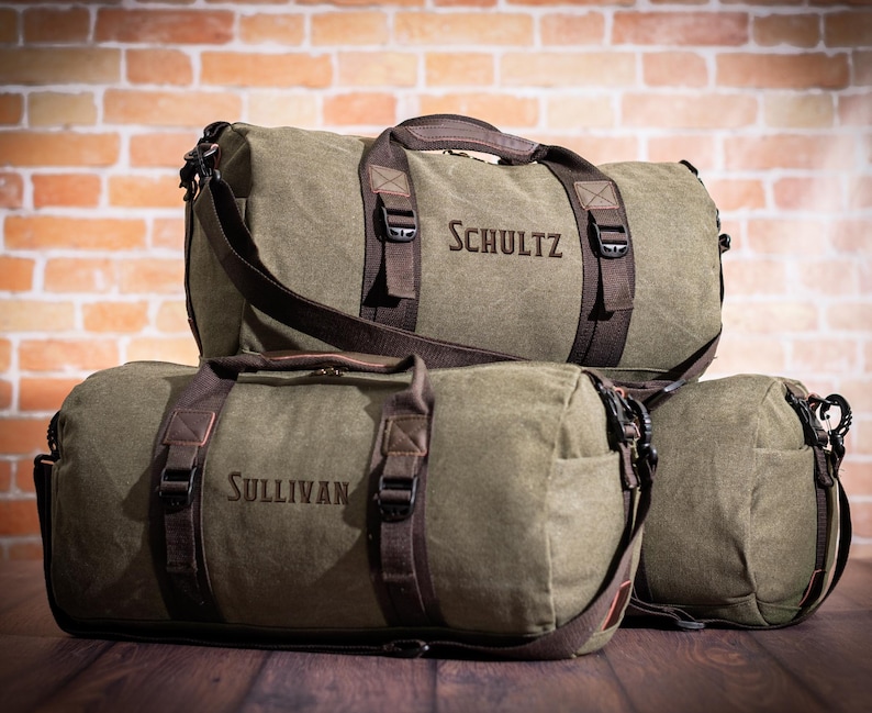 K&ouml;nnte beinhalten: Drei gr&uuml;ne Canvas-Reisetaschen mit braunen Lederakzenten und Riemen. Die Taschen sind mit den Namen "Schultz", "Sullivan" und einem dritten Namen personalisiert, der auf dem Bild nicht zu sehen ist.