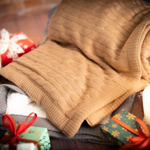 May include: A folded tan cable-knit blanket rests atop a gray blanket with a white interior. The scene includes lit candles in blue and white containers, and wrapped gifts with red ribbons. The items are arranged on a dark wooden surface, creating a cozy, festive atmosphere.