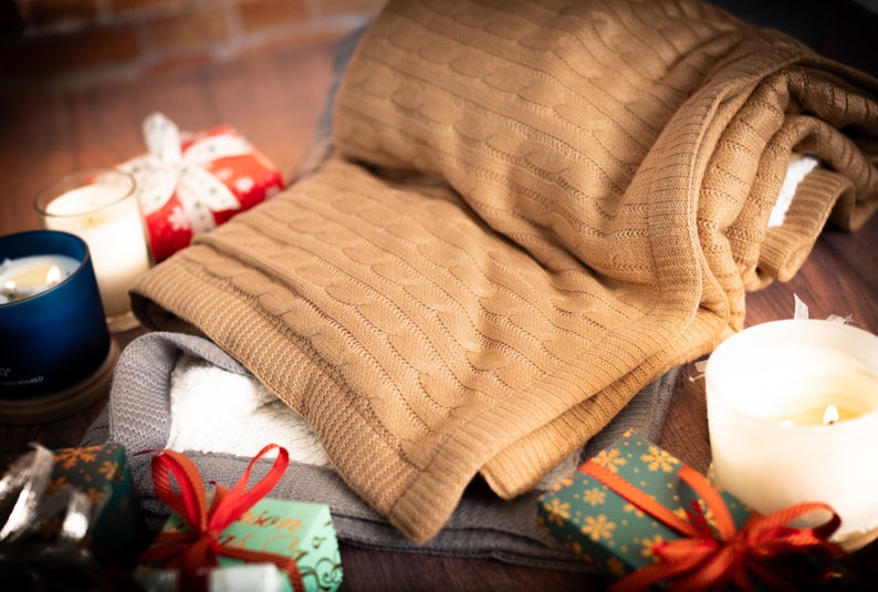 May include: A folded tan cable-knit blanket rests atop a gray blanket with a white interior. The scene includes lit candles in blue and white containers, and wrapped gifts with red ribbons. The items are arranged on a dark wooden surface, creating a cozy, festive atmosphere.