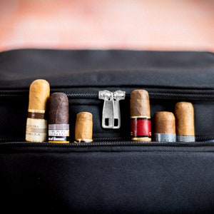 K&ouml;nnte beinhalten: Eine schwarze Reisetasche mit Rei&szlig;verschluss, die verschiedene Zigarren enth&auml;lt. Die Zigarren sind in einer Reihe angeordnet und haben unterschiedliche Marken und Gr&ouml;&szlig;en.