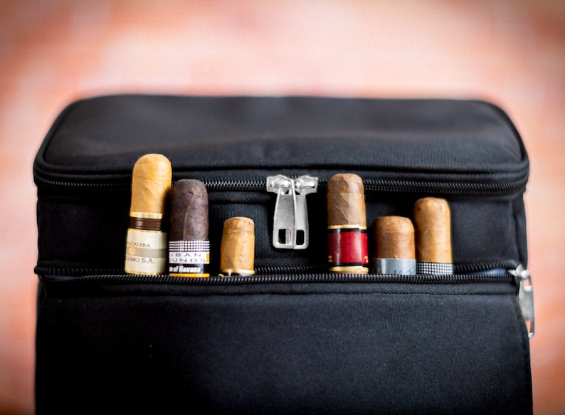 K&ouml;nnte beinhalten: Eine schwarze Reisetasche mit Rei&szlig;verschluss, die verschiedene Zigarren enth&auml;lt. Die Zigarren sind in einer Reihe angeordnet und haben unterschiedliche Marken und Gr&ouml;&szlig;en.