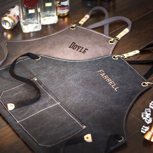 May include: Two personalised black canvas aprons with adjustable straps and a large pocket. The aprons are embroidered with the names "DOYLE" and "FARRELL".