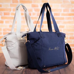 May include: Two tote bags, one light gray and one navy blue, with gold-colored hardware. The navy bag has the text "Karma BSN" in gold script. Both bags have shoulder straps and top handles, and are set against a brick wall.