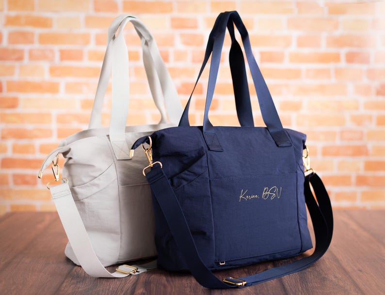 May include: Two tote bags, one light gray and one navy blue, with gold-colored hardware. The navy bag has the text "Karma BSN" in gold script. Both bags have shoulder straps and top handles, and are set against a brick wall.