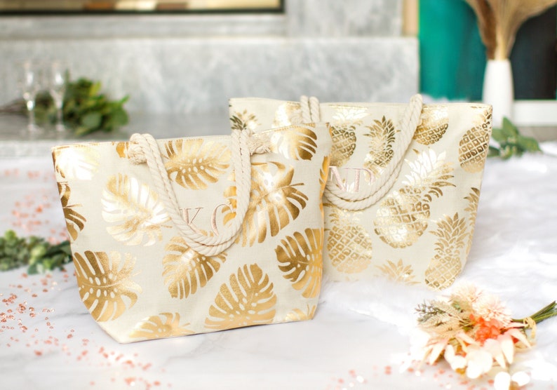 May include: Two white canvas tote bags with gold foil tropical leaf and pineapple designs. The bags have rope handles and are personalized with the initials "K" and "M".