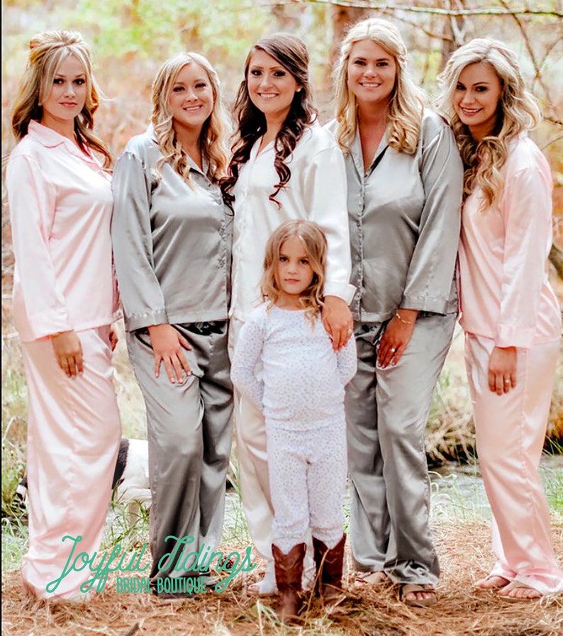 May include: Six women and a young girl wearing matching satin pajamas in pink and gray. The women are standing in a line, with the girl in front of them. They are all smiling and looking at the camera. The pajamas have a button-up top and long pants. The text "Joyful Tidings Bridal Boutique" is visible at the bottom of the image.