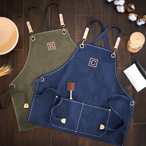 May include: Two canvas aprons with adjustable straps and pockets. One apron is olive green with the letter "R" embroidered on the front. The other apron is navy blue with the letter "C" embroidered on the front.