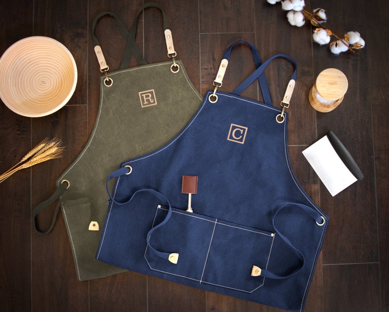 May include: Two canvas aprons with adjustable straps and pockets. One apron is olive green with the letter "R" embroidered on the front. The other apron is navy blue with the letter "C" embroidered on the front.