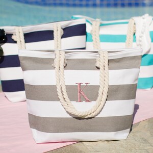 Puede incluir: Tres bolsas de lona a rayas con asas de cuerda. La bolsa en primer plano es gris y blanca con una letra K bordada en rosa. La bolsa del medio es azul y blanca. La bolsa del fondo es turquesa y blanca.