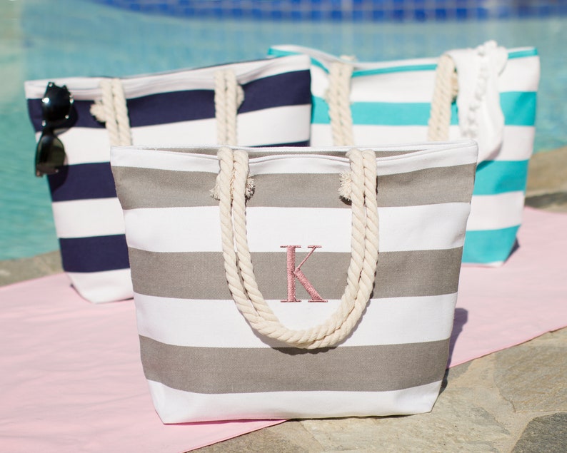 Puede incluir: Tres bolsas de lona a rayas con asas de cuerda. La bolsa en primer plano es gris y blanca con una letra K bordada en rosa. La bolsa del medio es azul y blanca. La bolsa del fondo es turquesa y blanca.