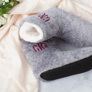 May include: A pair of gray fuzzy slippers with a white sherpa lining. The slippers have a black rubber sole and are embroidered with the initials "GRR" and "LM" in burgundy thread.