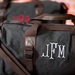 K&ouml;nnte beinhalten: Eine graue Canvas-Reisetasche mit braunen Lederakzenten und einem wei&szlig;en gestickten Monogramm "JFM".