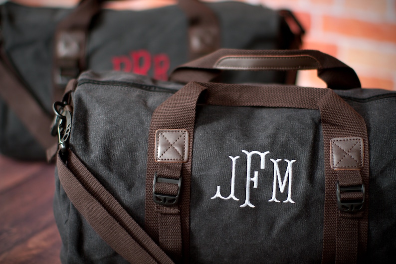 K&ouml;nnte beinhalten: Eine graue Canvas-Reisetasche mit braunen Lederakzenten und einem wei&szlig;en gestickten Monogramm "JFM".