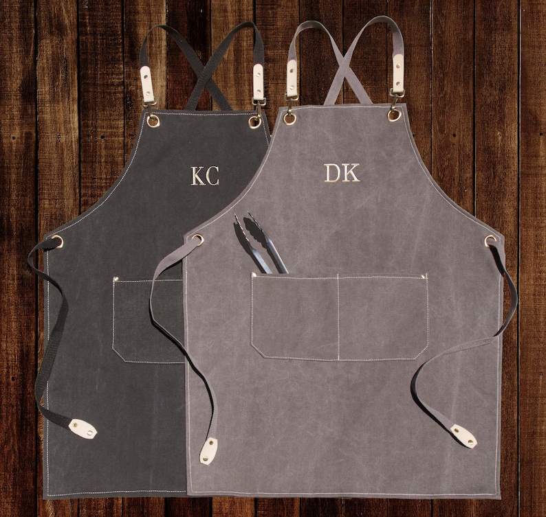 May include: Two canvas aprons with adjustable straps and a large pocket. The apron on the left is black with the initials "KC" embroidered on the front. The apron on the right is grey with the initials "DK" embroidered on the front.
