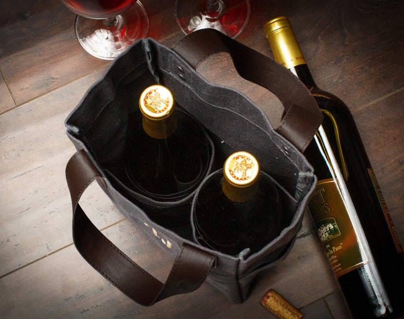 May include: A gray canvas wine tote bag with brown leather handles and two wine bottles inside. The tote bag has two separate compartments for the bottles. The bottles are both red wine and have gold foil caps.