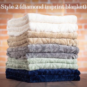 May include: A stack of eight plush blankets in various colors, including white, beige, gray, green, and blue. The blankets are folded and stacked on top of each other, creating a soft and inviting display. The text "Style 2 (diamond imprint blanket)" is visible at the top of the image.