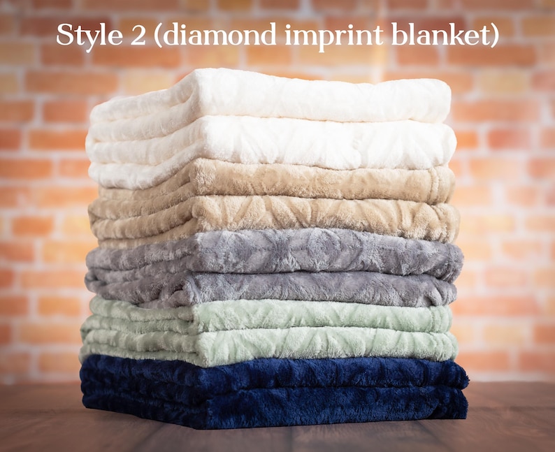 May include: A stack of eight plush blankets in various colors, including white, beige, gray, green, and blue. The blankets are folded and stacked on top of each other, creating a soft and inviting display. The text "Style 2 (diamond imprint blanket)" is visible at the top of the image.