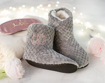 May include: A pair of gray fuzzy slippers with a black rubber sole. The slippers have a monogram embroidered on the top. The slippers are on a white surface with a pink eye mask and a pink flower.