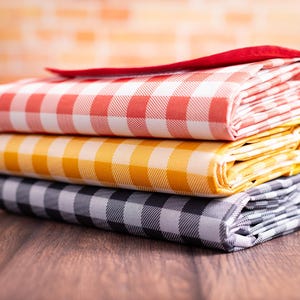 Puede incluir: Una pila de tres manteles a cuadros plegados en rojo, amarillo y negro. El mantel rojo est&aacute; en la parte superior, con un borde rojo liso. Los manteles amarillo y negro est&aacute;n debajo. Los manteles est&aacute;n apilados sobre una superficie de madera.