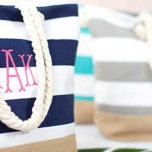 May include: Canvas tote bags with navy blue and white stripes, beige bottoms, and rope handles. One bag has pink embroidered initials. Other bags in the background have similar designs in different colors. A stylish and practical accessory.