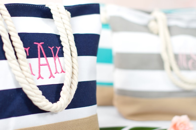 May include: Canvas tote bags with navy blue and white stripes, beige bottoms, and rope handles. One bag has pink embroidered initials. Other bags in the background have similar designs in different colors. A stylish and practical accessory.