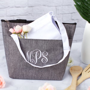 May include: A gray insulated tote bag with a white zipper and a white handle. The bag has a white monogram that says "MPS" on the front. The bag is sitting on a white surface with pink flowers and wooden kitchen utensils.