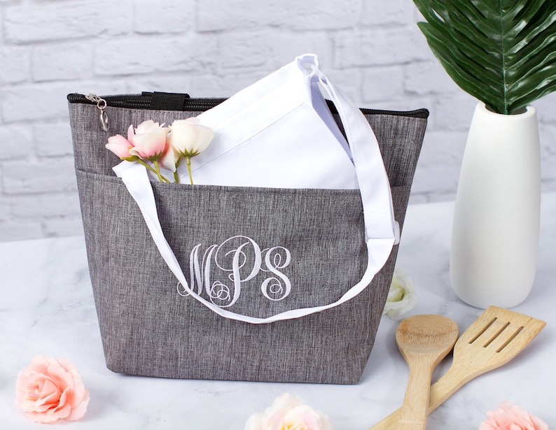 May include: A gray insulated tote bag with a white zipper and a white handle. The bag has a white monogram that says "MPS" on the front. The bag is sitting on a white surface with pink flowers and wooden kitchen utensils.