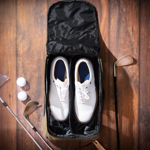 May include: A black travel bag with two white golf shoes inside. The bag has a zipper closure and a handle. There are two golf clubs and three golf balls on the wooden floor.