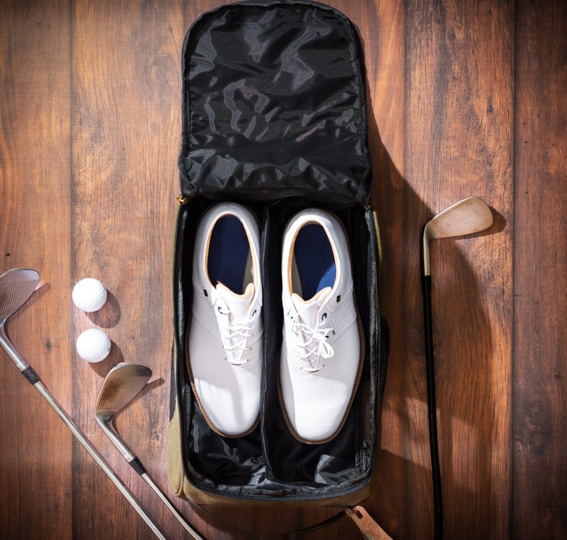 May include: A black travel bag with two white golf shoes inside. The bag has a zipper closure and a handle. There are two golf clubs and three golf balls on the wooden floor.