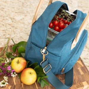 May include: Teal corduroy lunch bag with a gold zipper and hardware, tan leather handles, and a detachable shoulder strap. The bag is open, revealing a glass container filled with red cherry tomatoes. Two apples and flowers are next to the bag.