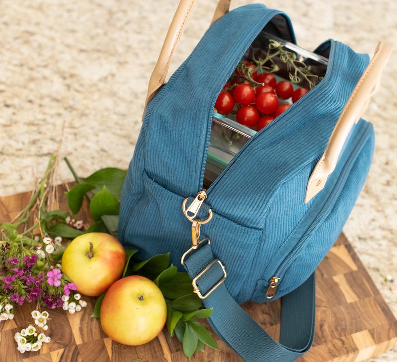 May include: Teal corduroy lunch bag with a gold zipper and hardware, tan leather handles, and a detachable shoulder strap. The bag is open, revealing a glass container filled with red cherry tomatoes. Two apples and flowers are next to the bag.