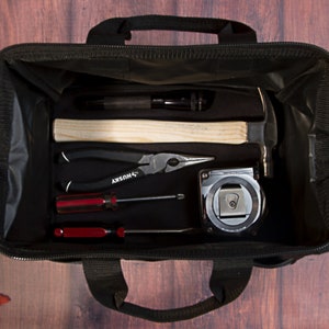 K&ouml;nnte beinhalten: Eine schwarze Werkzeugtasche mit Rei&szlig;verschluss, die einen Hammer, eine Zange, Schraubendreher und ein Ma&szlig;band enth&auml;lt. Die Tasche ist offen und die Werkzeuge sind sichtbar.