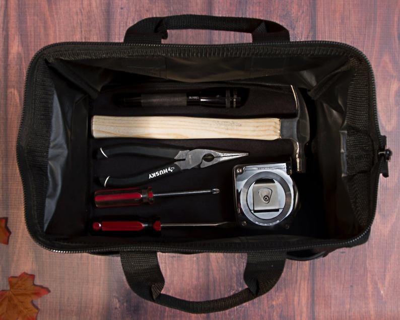 K&ouml;nnte beinhalten: Eine schwarze Werkzeugtasche mit Rei&szlig;verschluss, die einen Hammer, eine Zange, Schraubendreher und ein Ma&szlig;band enth&auml;lt. Die Tasche ist offen und die Werkzeuge sind sichtbar.