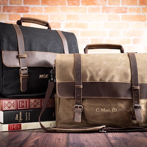 May include: A black canvas messenger bag with brown leather straps and the initials MGK, and a tan canvas messenger bag with brown leather straps and the text C. Marc, JD. Both bags feature top handles, adjustable shoulder straps, and metal buckles.