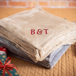 May include: A beige and gray fleece throw blanket with the monogram "B&T" embroidered in red on the top layer.