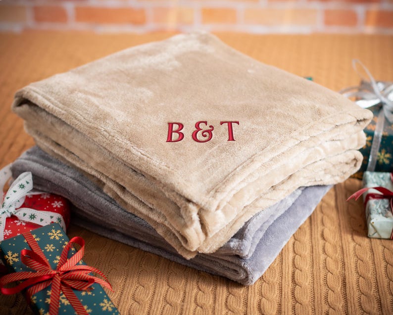 May include: A beige and gray fleece throw blanket with the monogram "B&T" embroidered in red on the top layer.