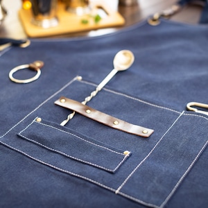 May include: A dark blue denim apron with a brown leather strap and a silver metal bar spoon. The apron has a large pocket with a white stitching detail.