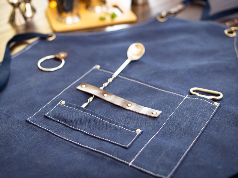 May include: A dark blue denim apron with a brown leather strap and a silver metal bar spoon. The apron has a large pocket with a white stitching detail.