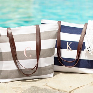 May include: Two striped canvas tote bags with brown leather handles. The bag on the left is gray and white with the letter "C" embroidered on it. The bag on the right is navy blue and white with the letter "K" embroidered on it.