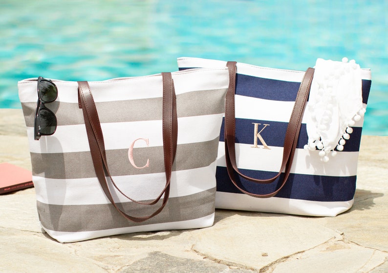 May include: Two striped canvas tote bags with brown leather handles. The bag on the left is gray and white with the letter "C" embroidered on it. The bag on the right is navy blue and white with the letter "K" embroidered on it.
